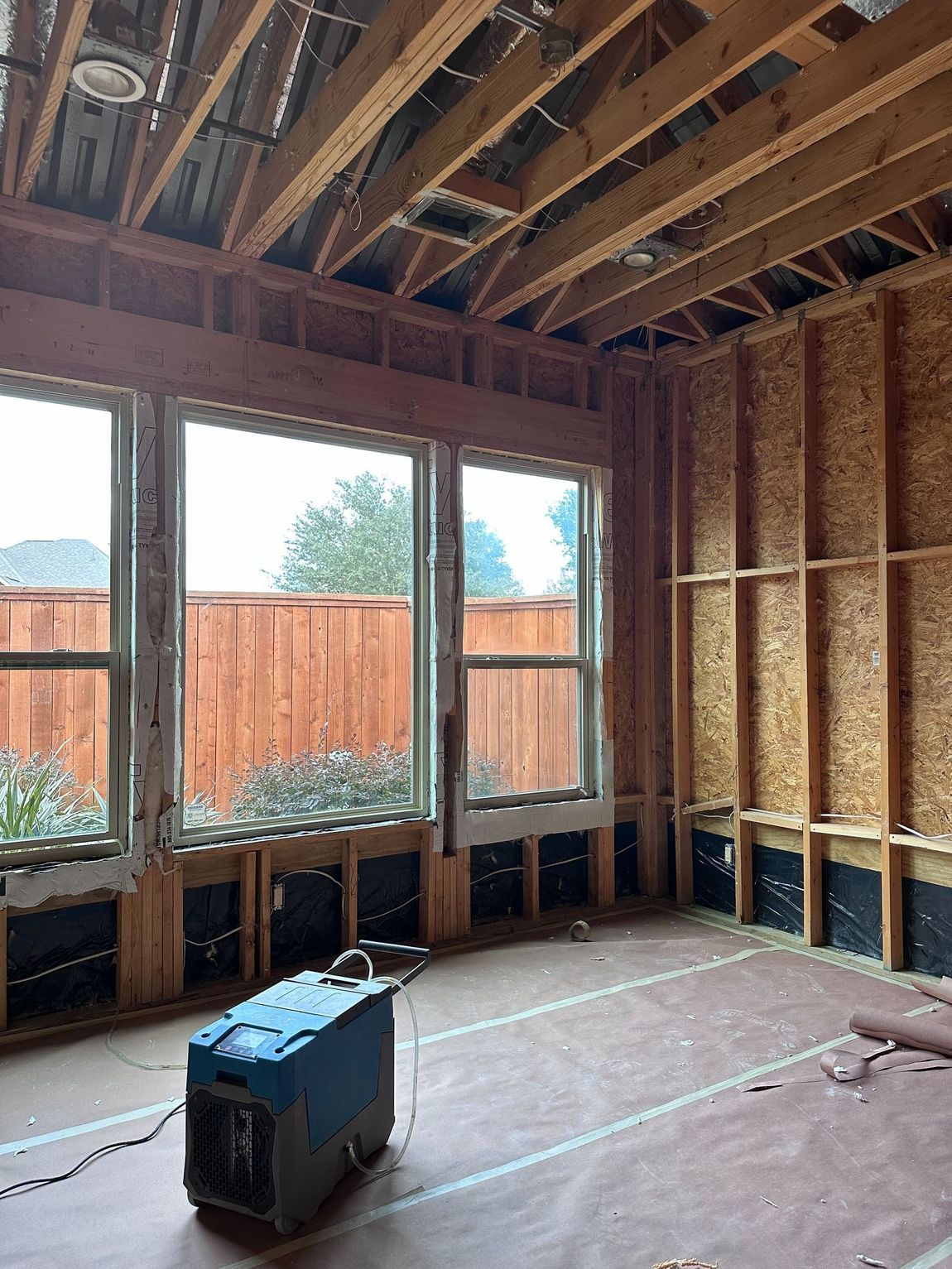 Interior of a room under construction with windows, exposed framing, and a dehumidifier on the floor.