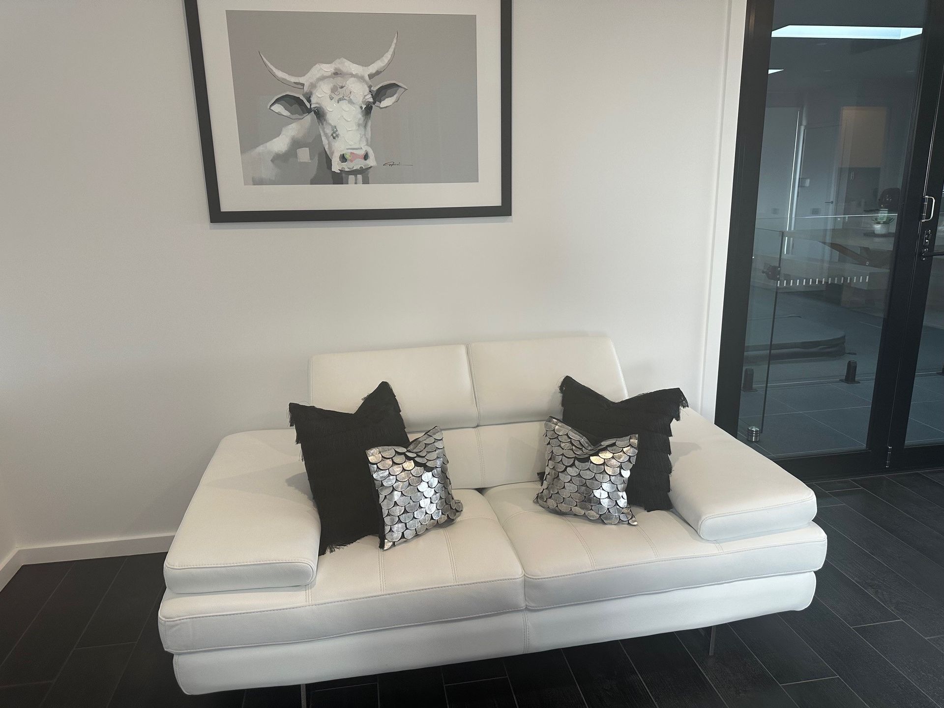 A white couch with black pillows and a picture of a cow on the wall above it