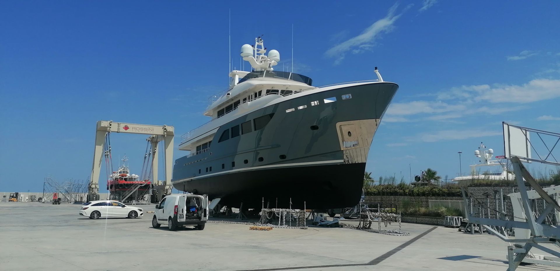 Un grande yacht in un bacino di carenaggio, grigio e bianco con lo scafo nero, sotto un cielo azzurro.