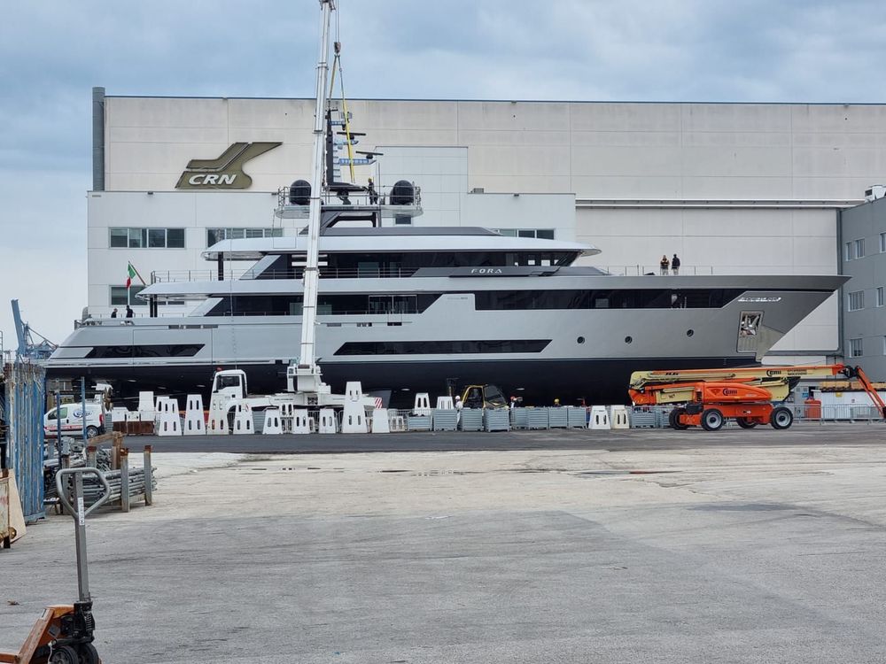 Un grande yacht bianco attraccato in un porto, con edifici e una collina sullo sfondo, sotto un cielo azzurro.