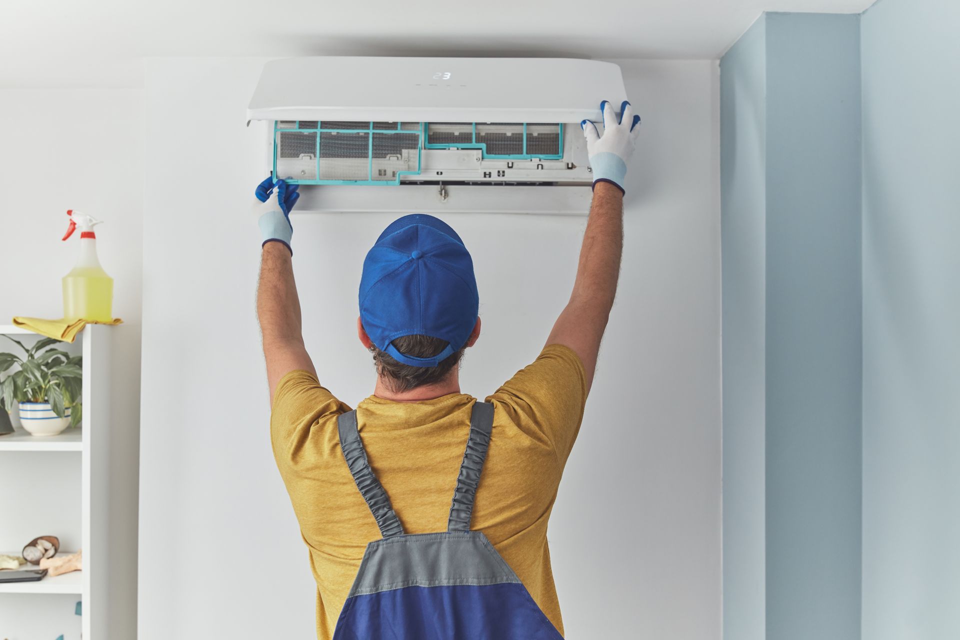 A technician wearing a blue hat and overalls works on the filters of a wall-mounted air conditioner.