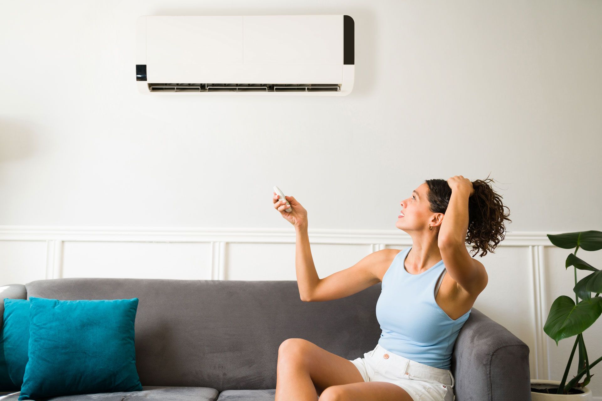 Woman sitting on a sofa, using a remote in a bright living room with a wall-mounted air conditioner.