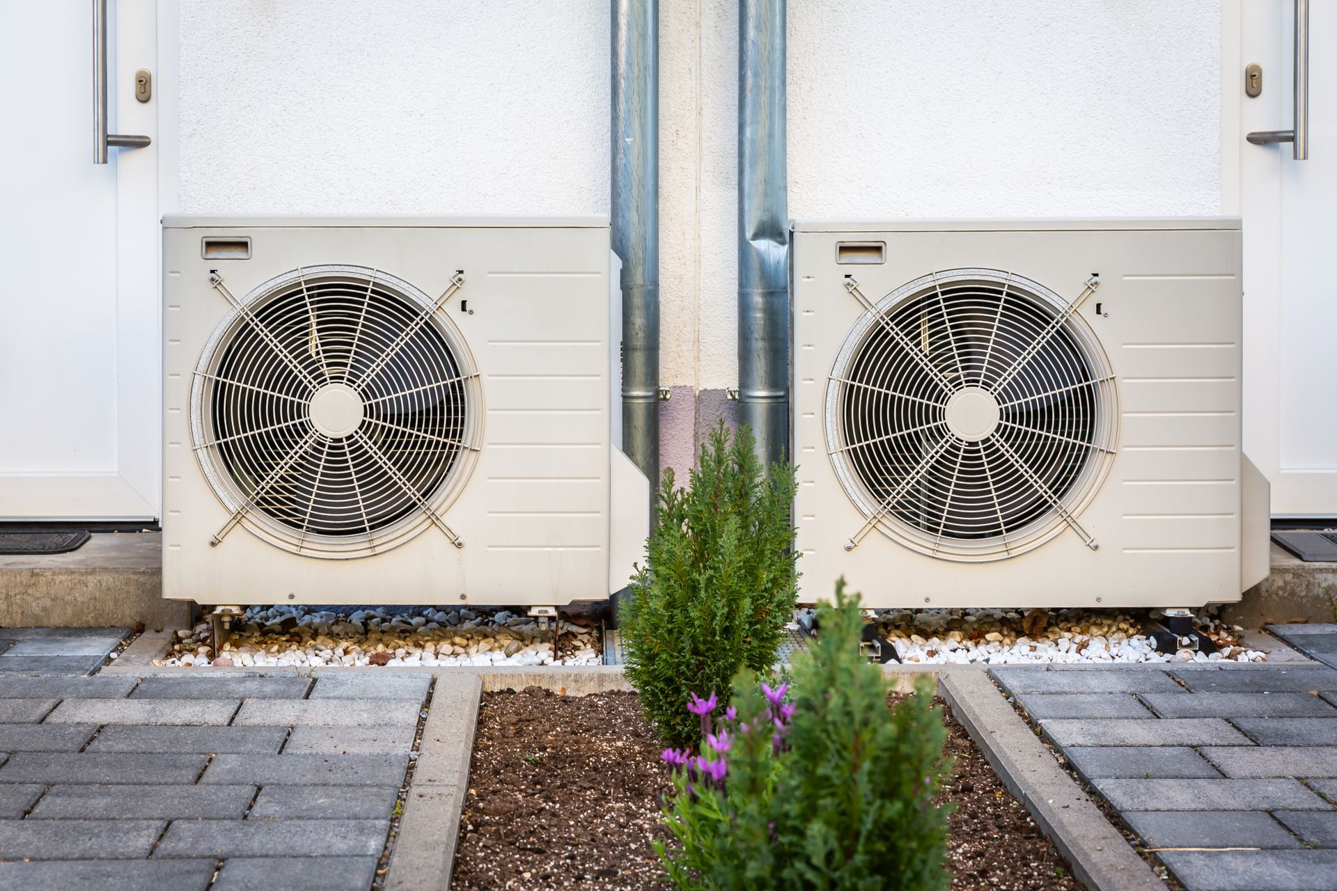 Two outdoor AC units beside a building, separated by a reflective metal column and landscaped path.
