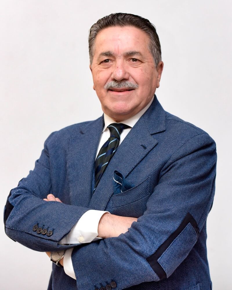 Ritratto di un uomo in abito blu con le braccia incrociate, in piedi davanti a uno sfondo chiaro e uniforme.