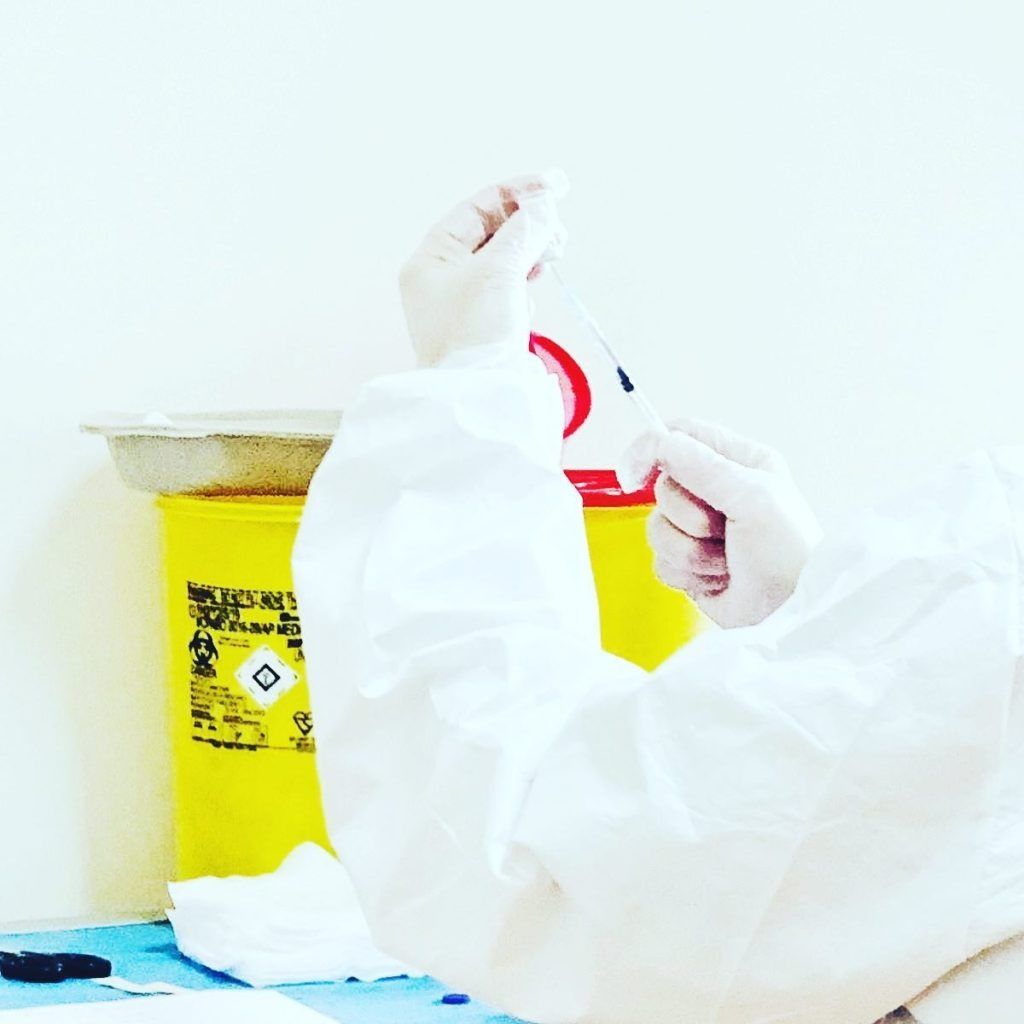 Una persona con i guanti maneggia una tuta protettiva bianca accanto a un bidone giallo per rifiuti chimici.