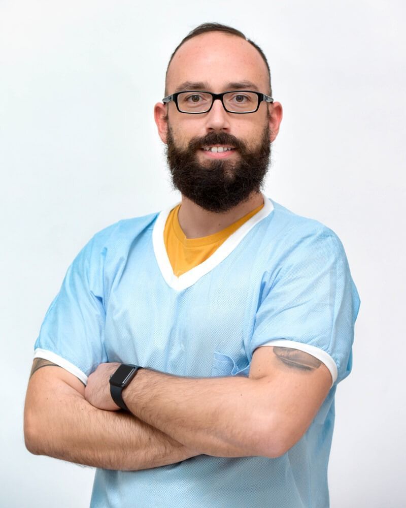 Persona sorridente con occhiali e barba, braccia incrociate, che indossa una camicia azzurra su sfondo bianco.