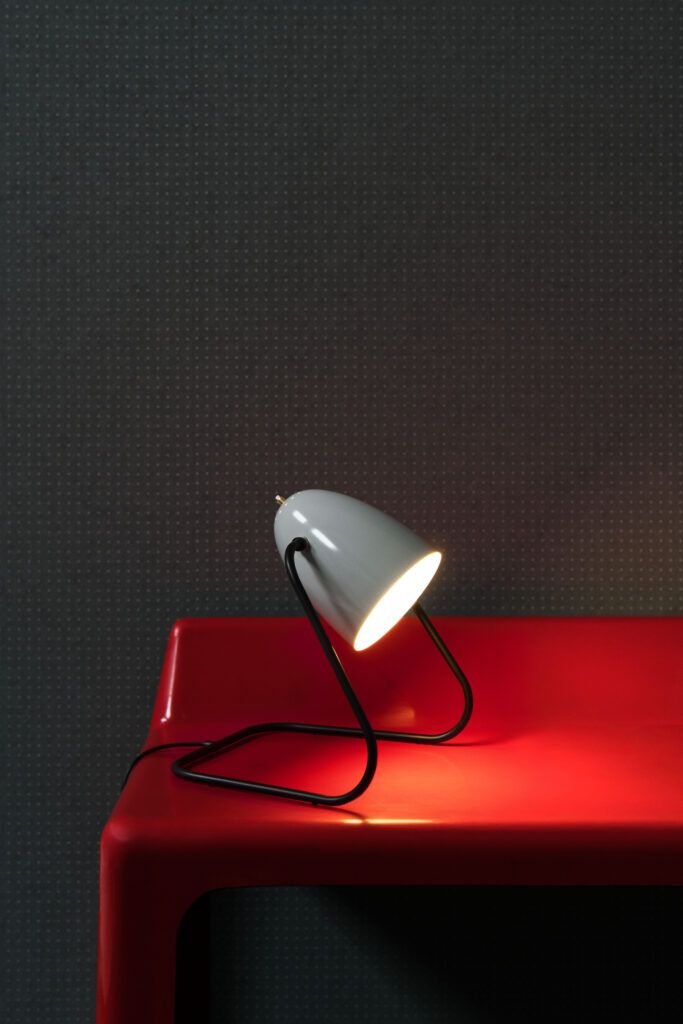 Hvit skrivebordslampe på et rødt bord mot en prikkete grå vegg.