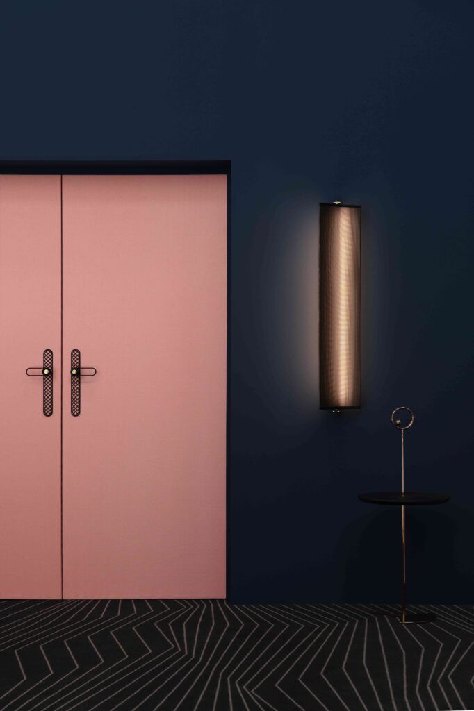 Rosa doble dører, mørkeblå vegg, sylindrisk vegglampe, lite bord, mønstret teppe.
