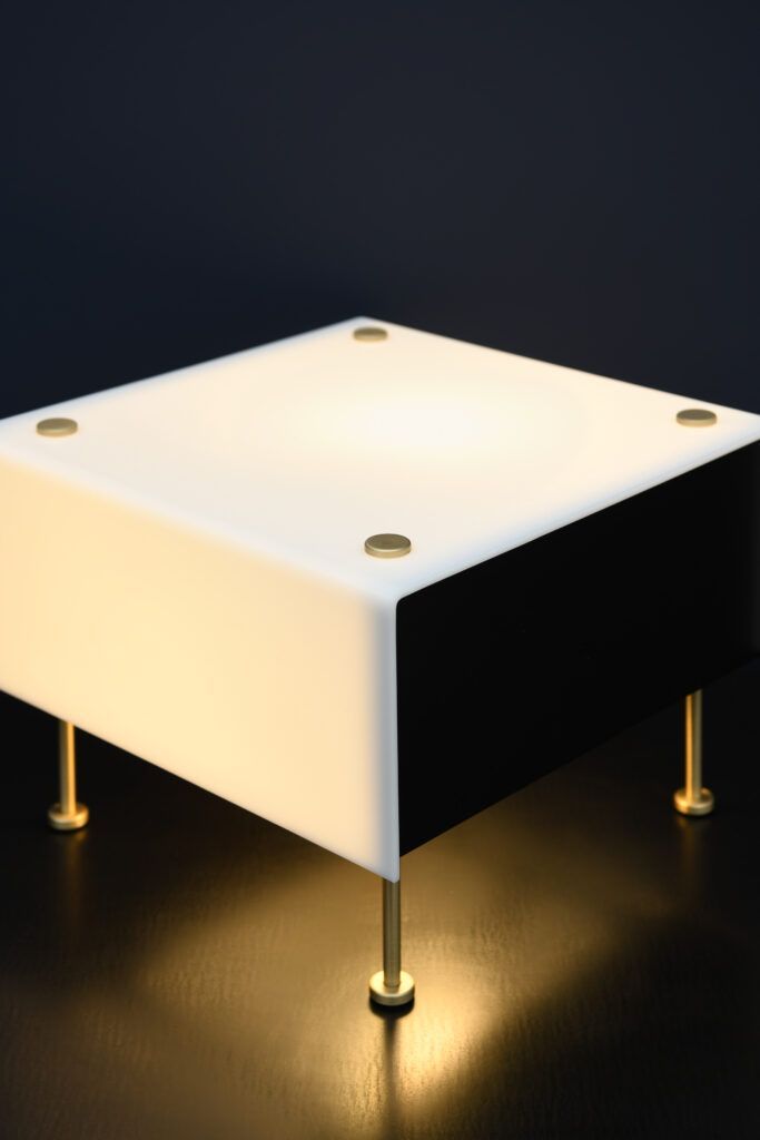 En firkantet bordlampe med svart og hvitt design, gullfargede ben og en lysende topp.