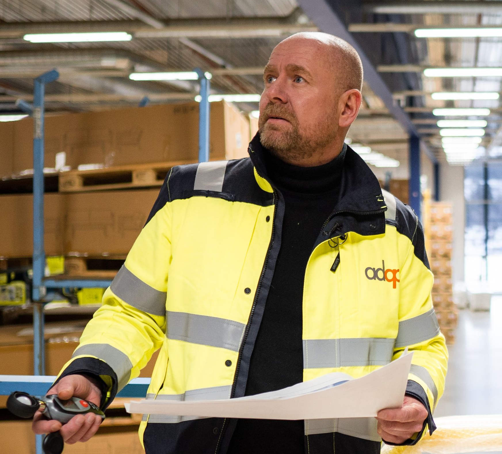 Mann i gul arbeidsjakke på et lager, holder papirer og ser opp.