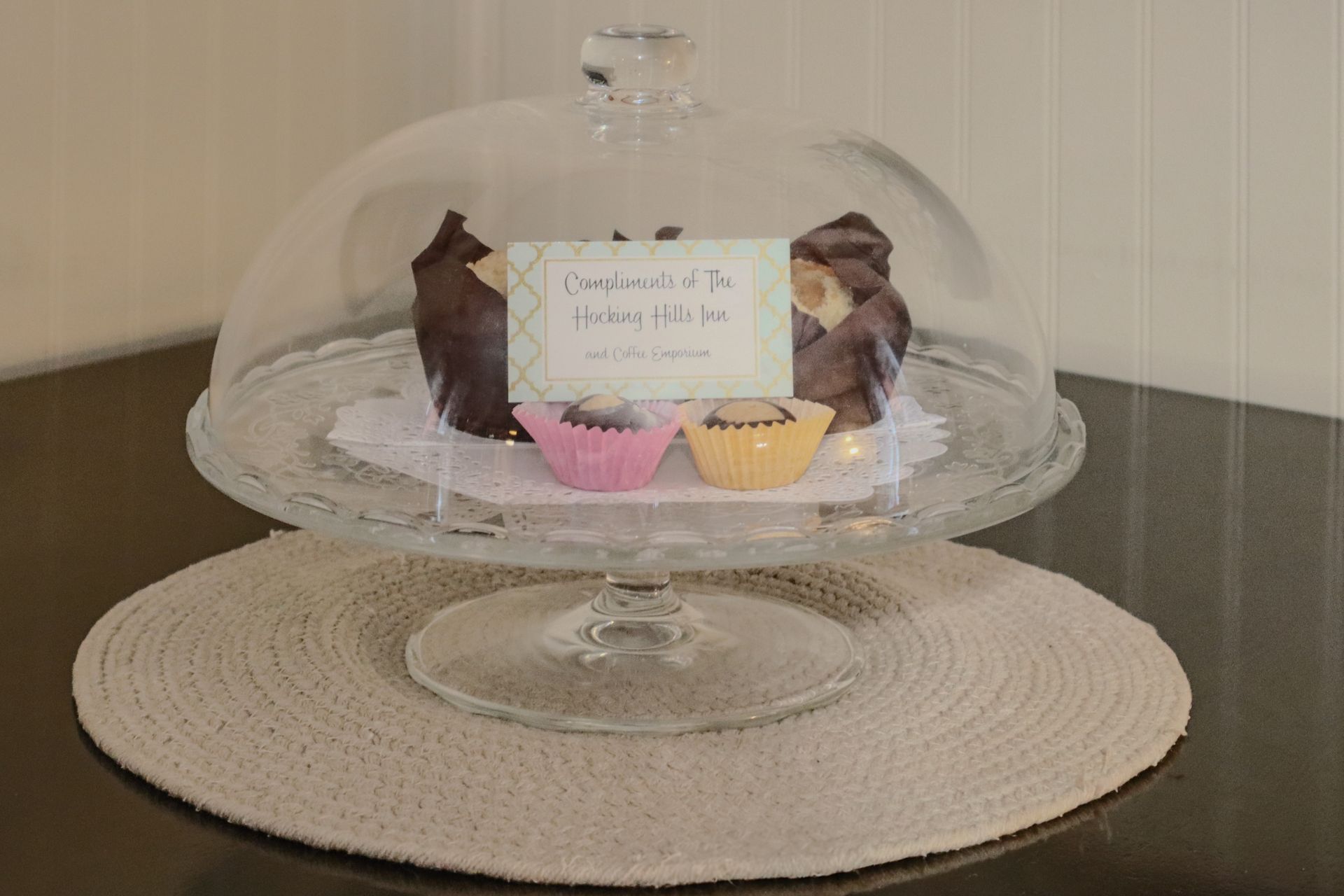 Cakes displayed under a glass dome on a white doily and a round placemat. Includes a sign and pink and yellow cupcakes.