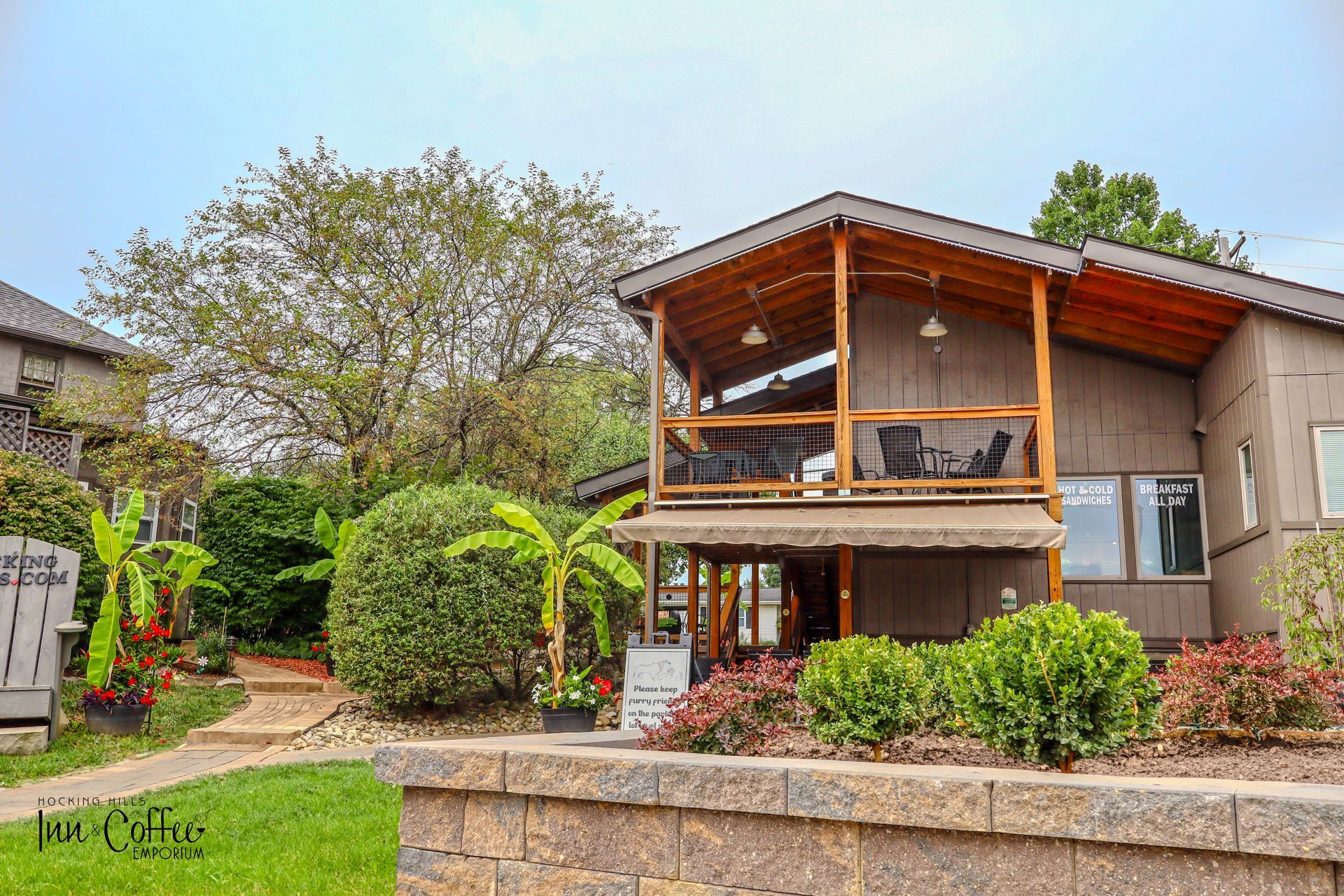 House with a brown exterior and a covered balcony, surrounded by landscaping, a brick walkway, and a retaining wall.