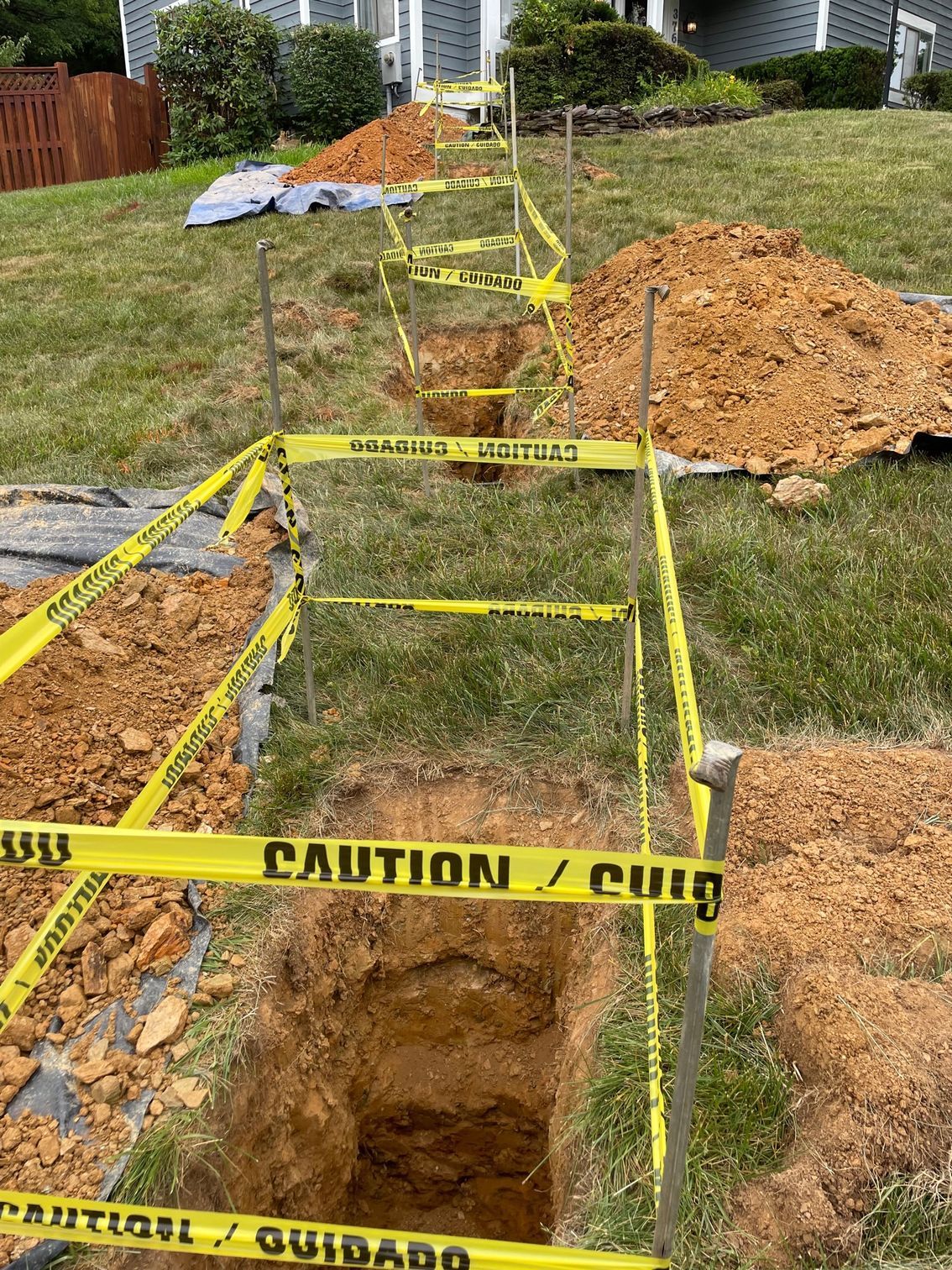 Caution tape around a long open trench in a grassy yard with dirt piles nearby.