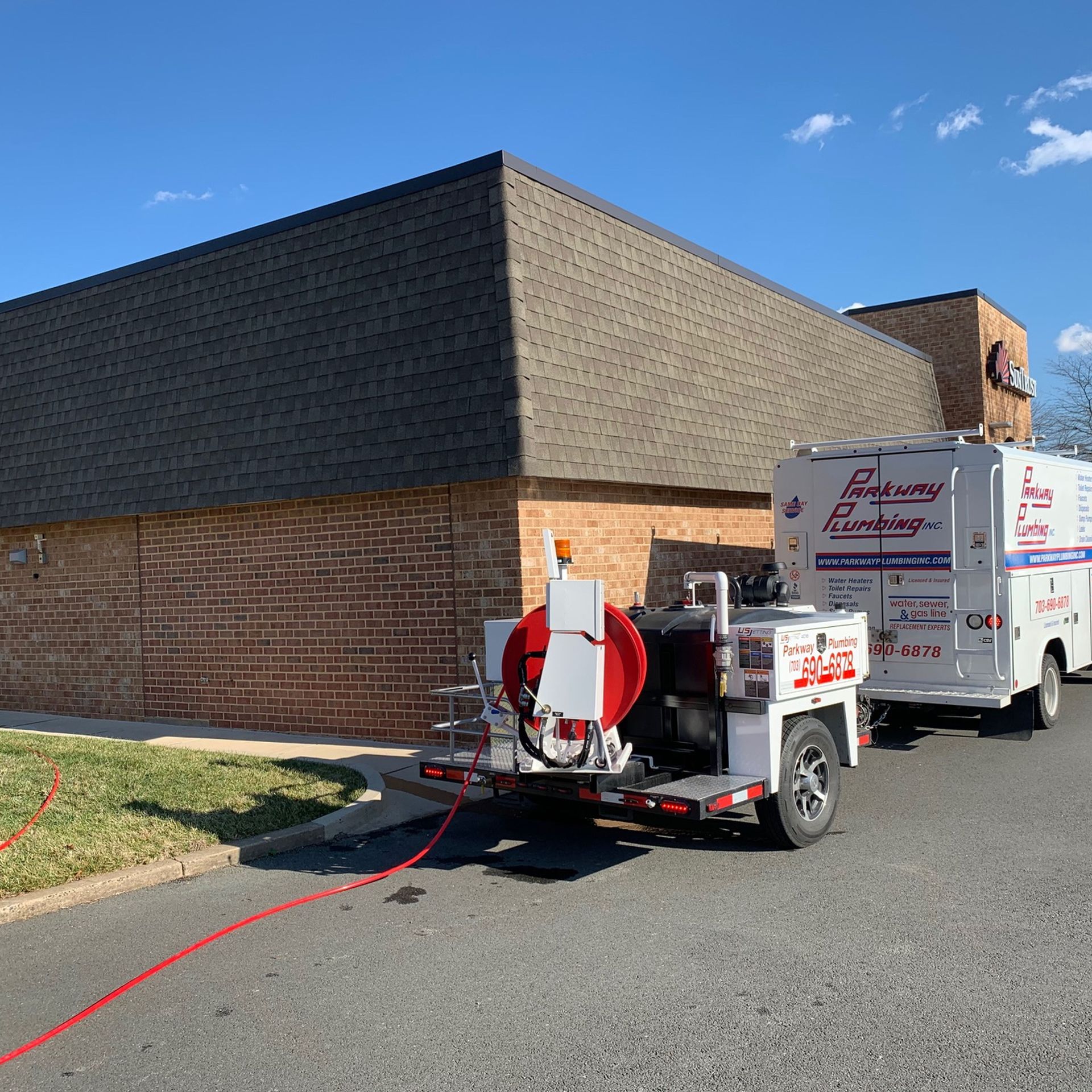 Utility trailer and white service truck parked beside a brick building, with red hose equipment attached.