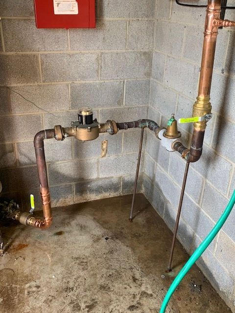 Plumbing pipes and valves along a cinder block wall in a utility room, with a green hose on the floor.