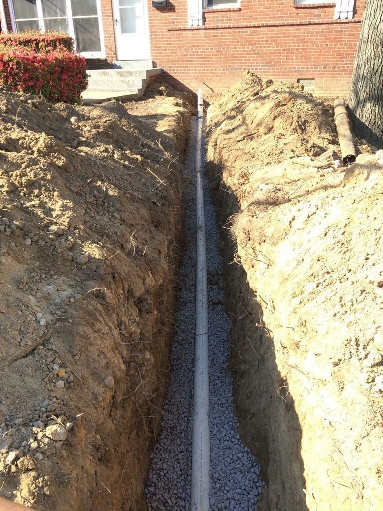 Narrow trench with gravel and a gray pipe running toward a brick house foundation
