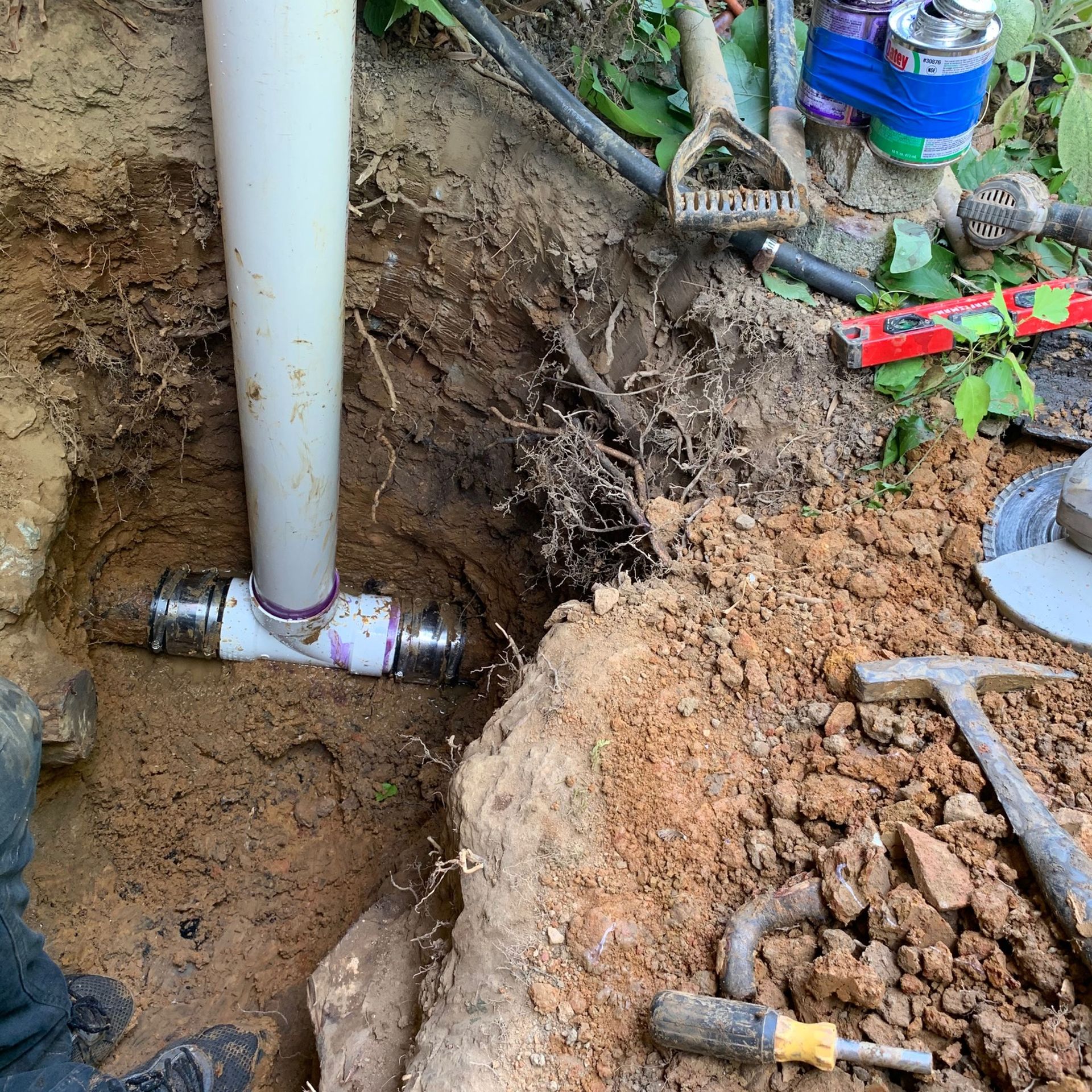 Excavated trench with a white pipe and PVC fittings, surrounded by dirt and tools.