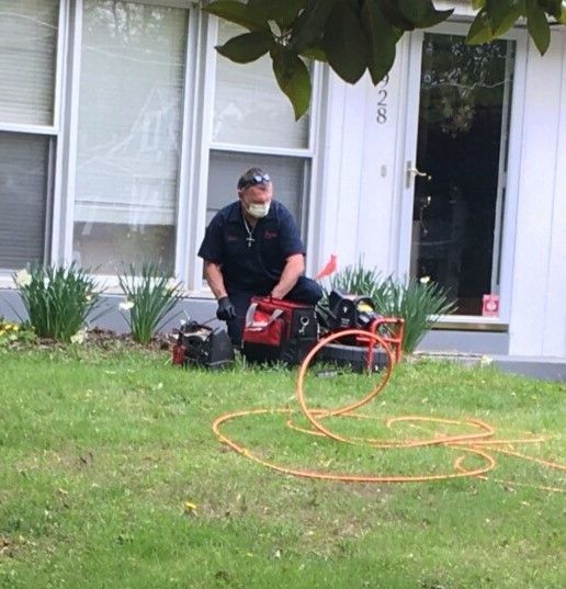 Worker kneeling on lawn beside red power tool and orange hose outside a house