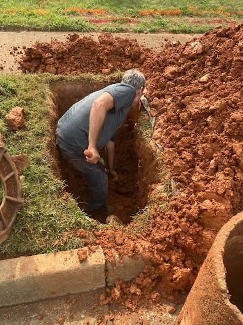 Person crouching in a muddy dirt hole beside a garden, with fresh soil piled around it.