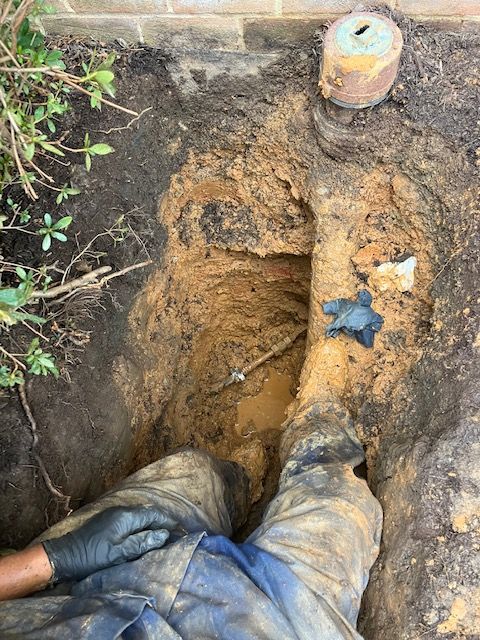 Worker in muddy trench with exposed pipe beside a brick wall and soil debris.