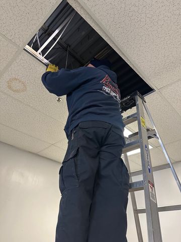 Person on a ladder installing ceiling panels in a room with an open ceiling grid.