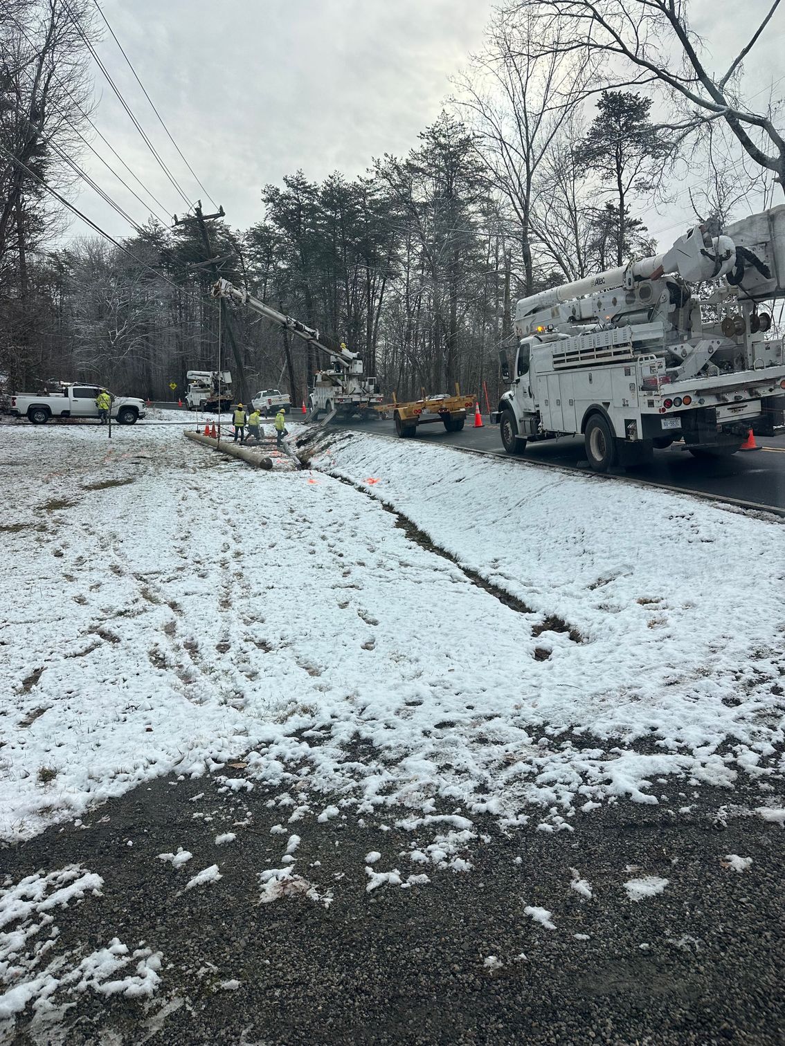 Snowy roadside with utility trucks and workers clearing fallen trees after storm damage.