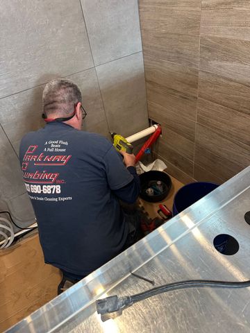 Person kneeling in a tiled shower stall, working on plumbing with red and yellow tools.