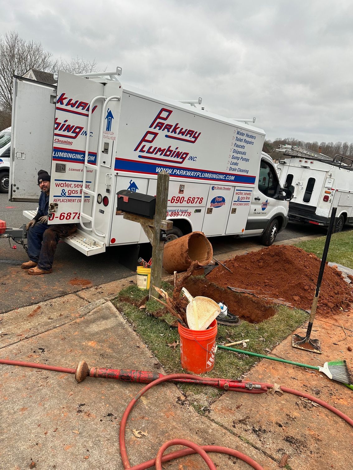 Workers at a plumbing truck digging a trench beside a house, with a bucket and hoses on the ground.