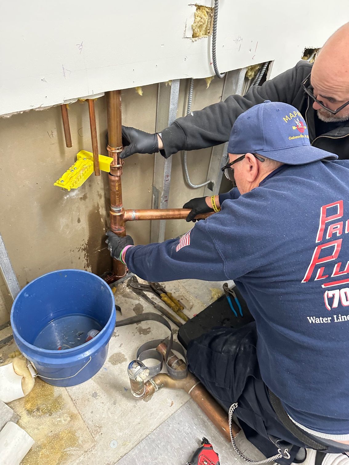 Two workers repairing plumbing under a wall, with tools and a blue bucket on a concrete floor