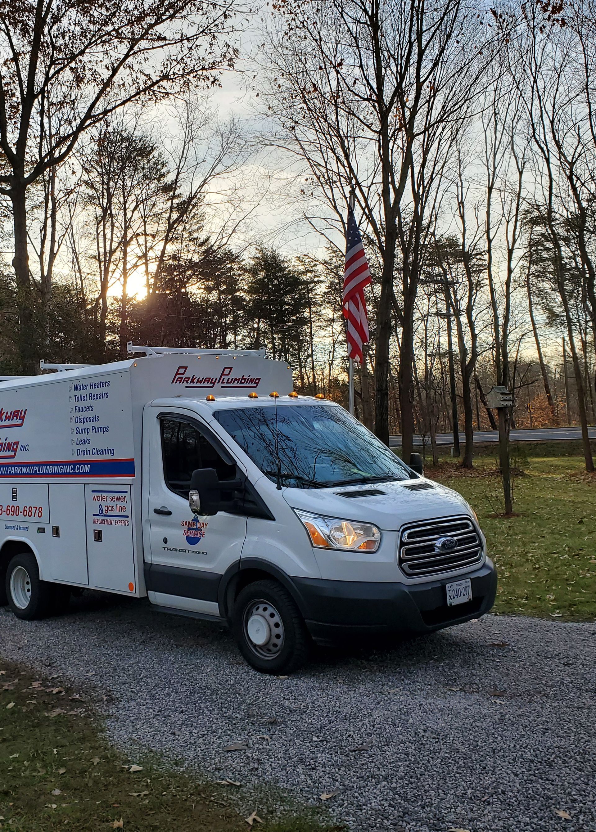 White utility truck parked on gravel at sunset, with an American flag and bare trees behind it