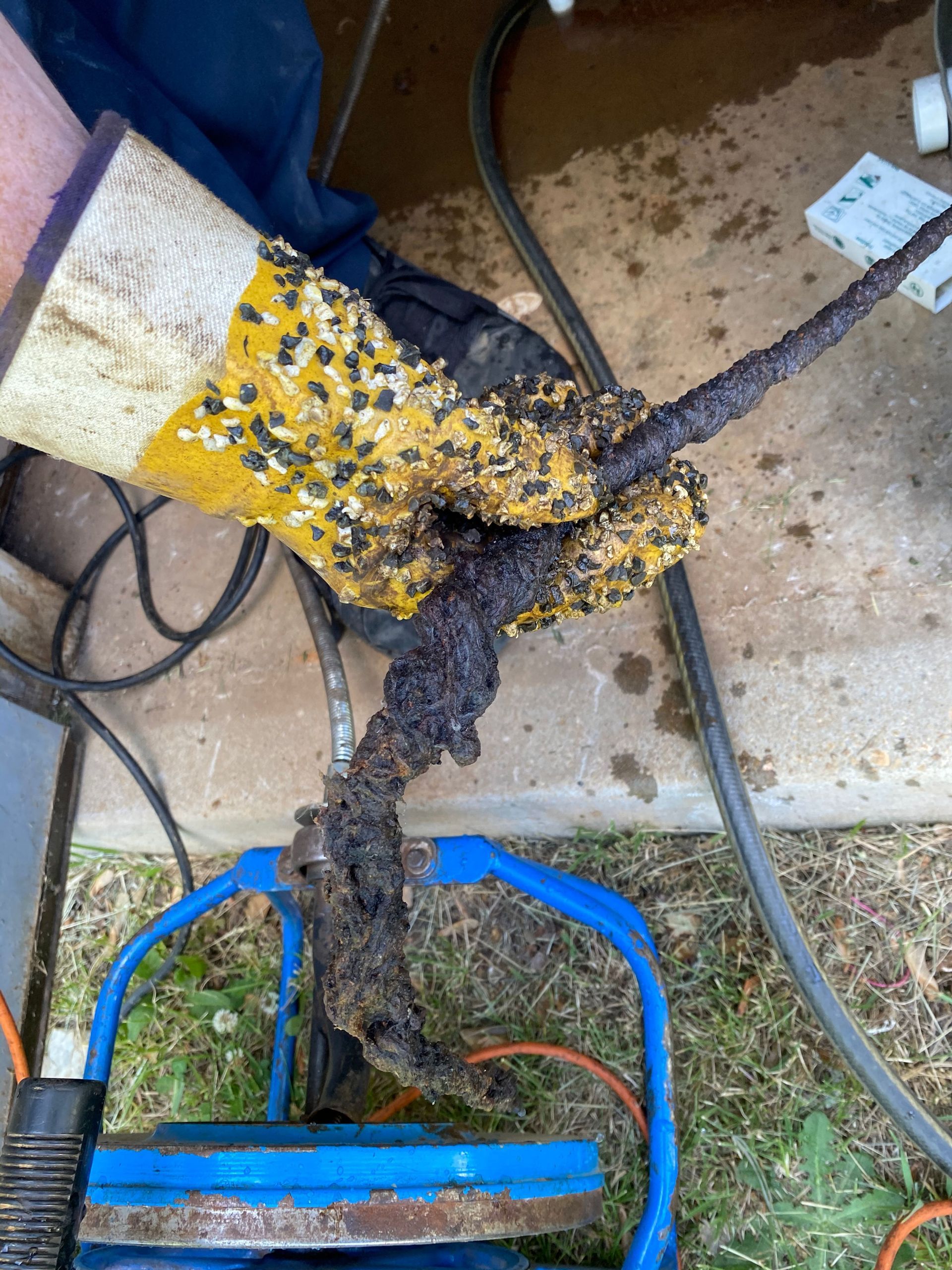 Yellow and black equipment wrapped in debris, with a hose and wires on a muddy outdoor ground.