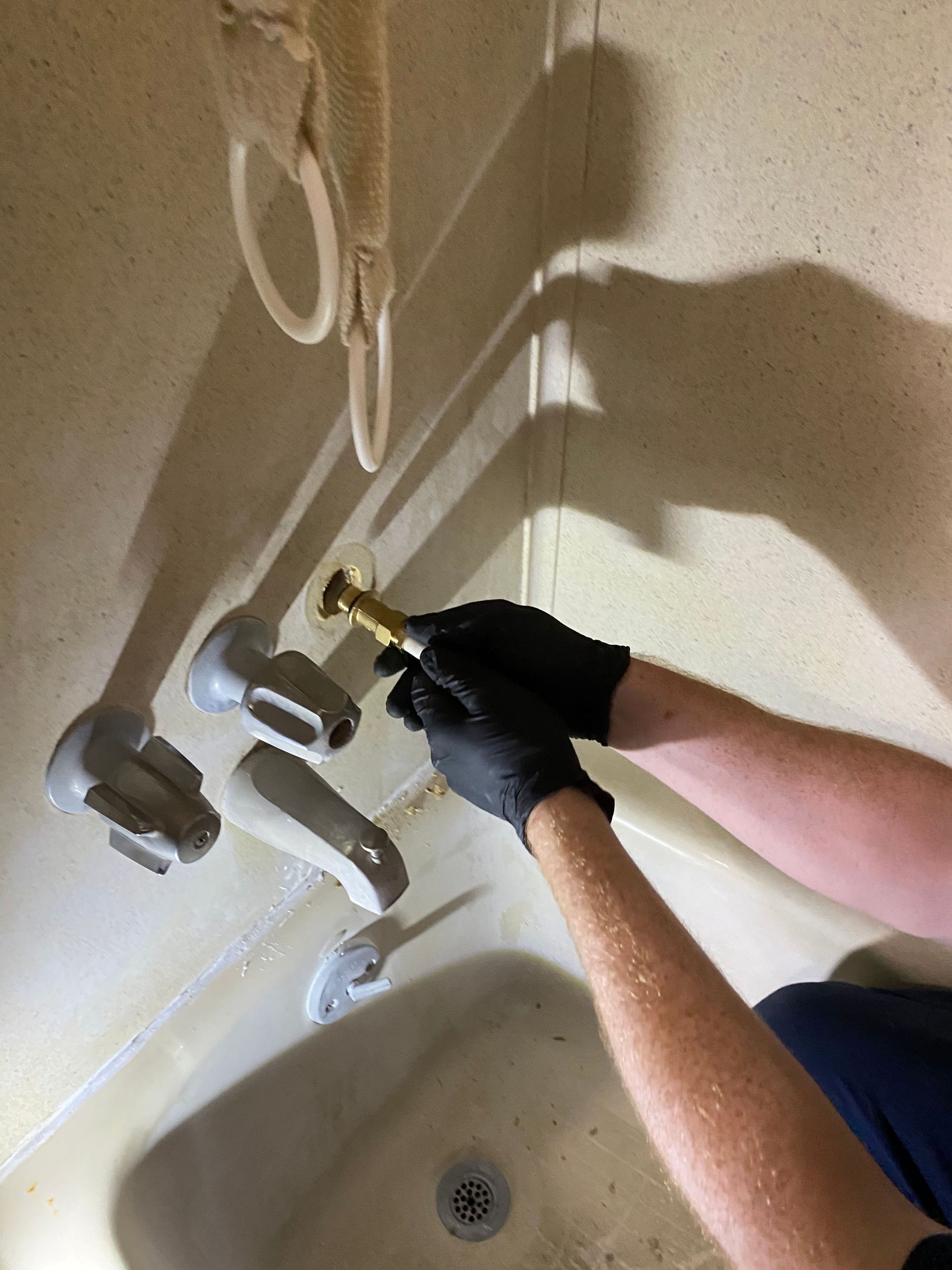Gloved person adjusting a shower faucet with exposed plumbing above a bathtub
