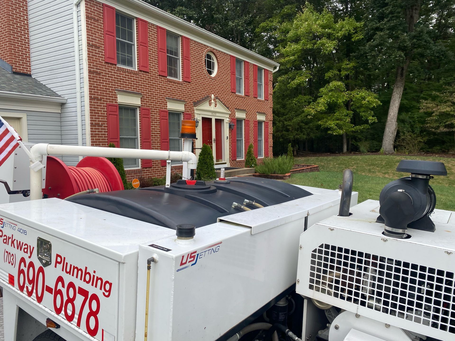 Plumbing service truck parked outside a brick house with red shutters and trees nearby