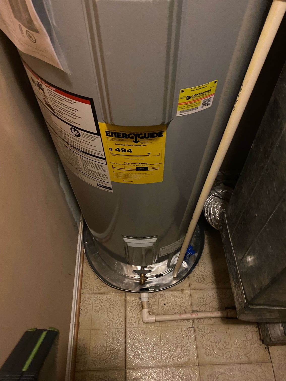 Water heater in a narrow utility closet with exposed pipes and tiled floor.