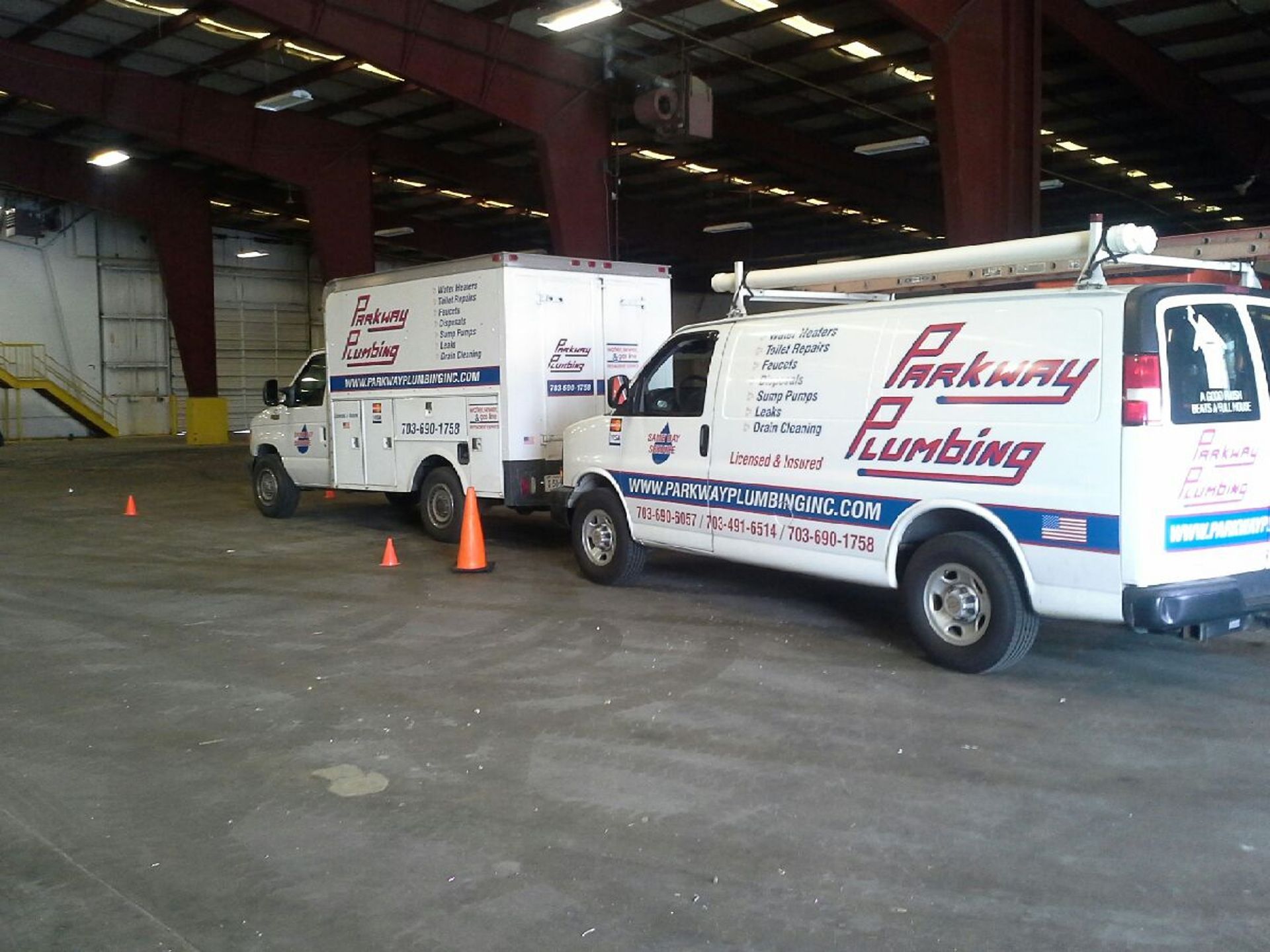 Two white plumbing service vans parked inside a warehouse, with orange cones nearby.
