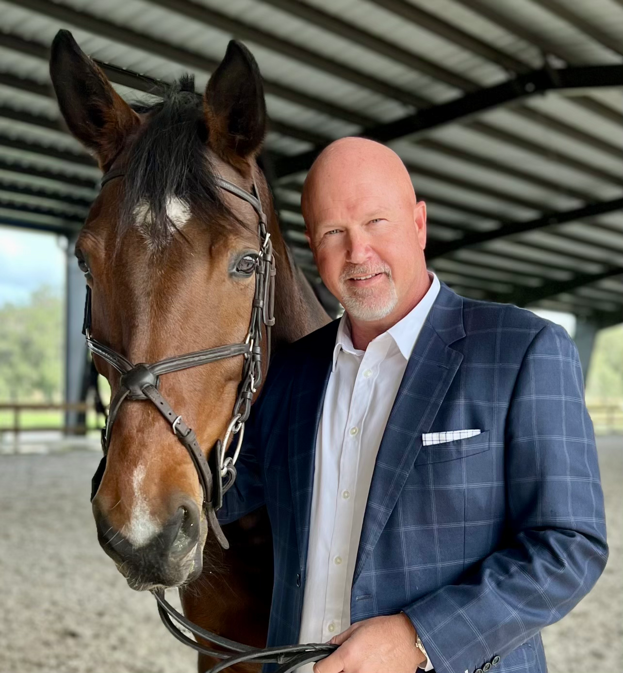 A person in a blue blazer stands beside a brown horse wearing a bridle inside an indoor riding arena.