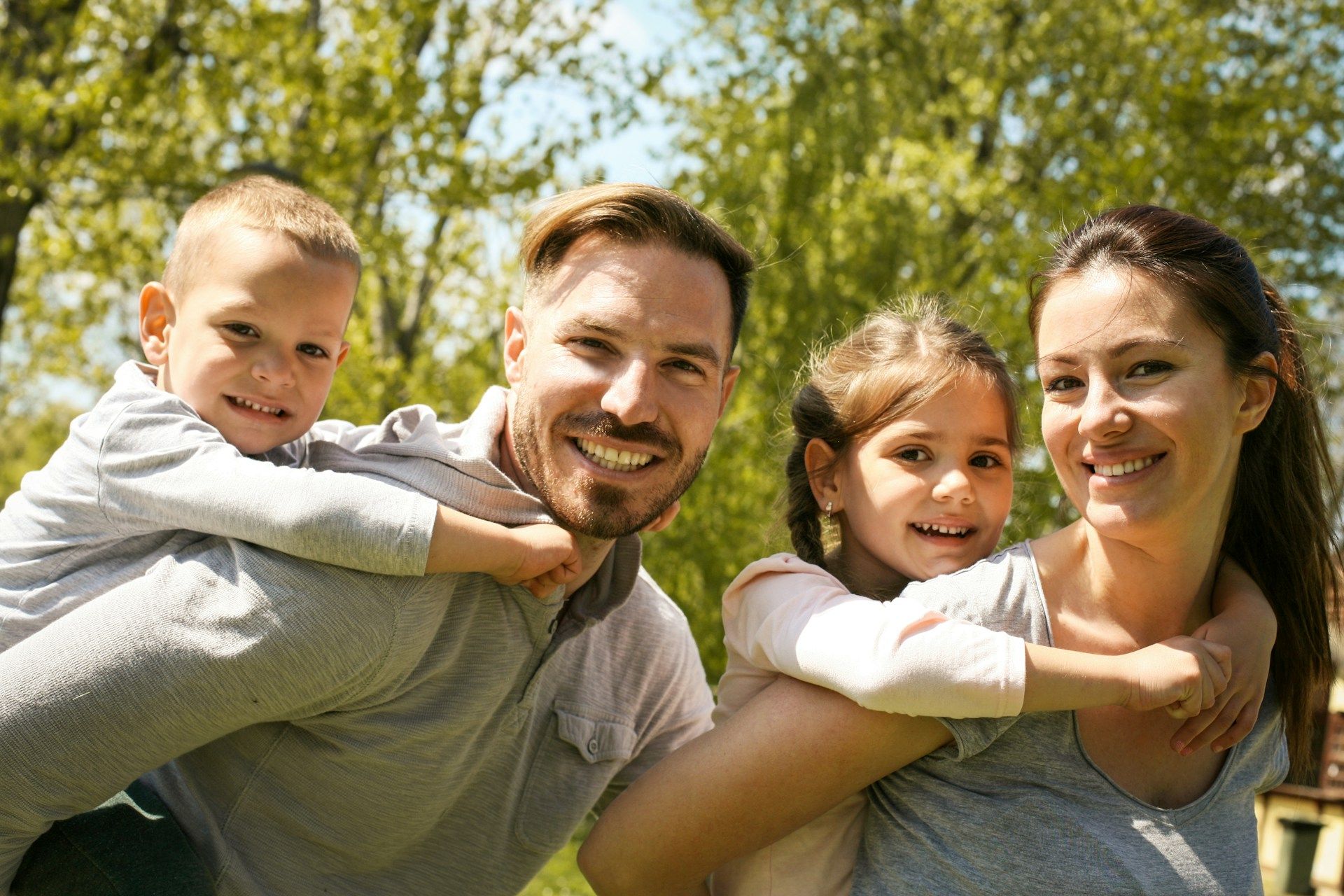 A parent carries a young child on their back on each side while standing outdoors in a sunlit, leafy environment.