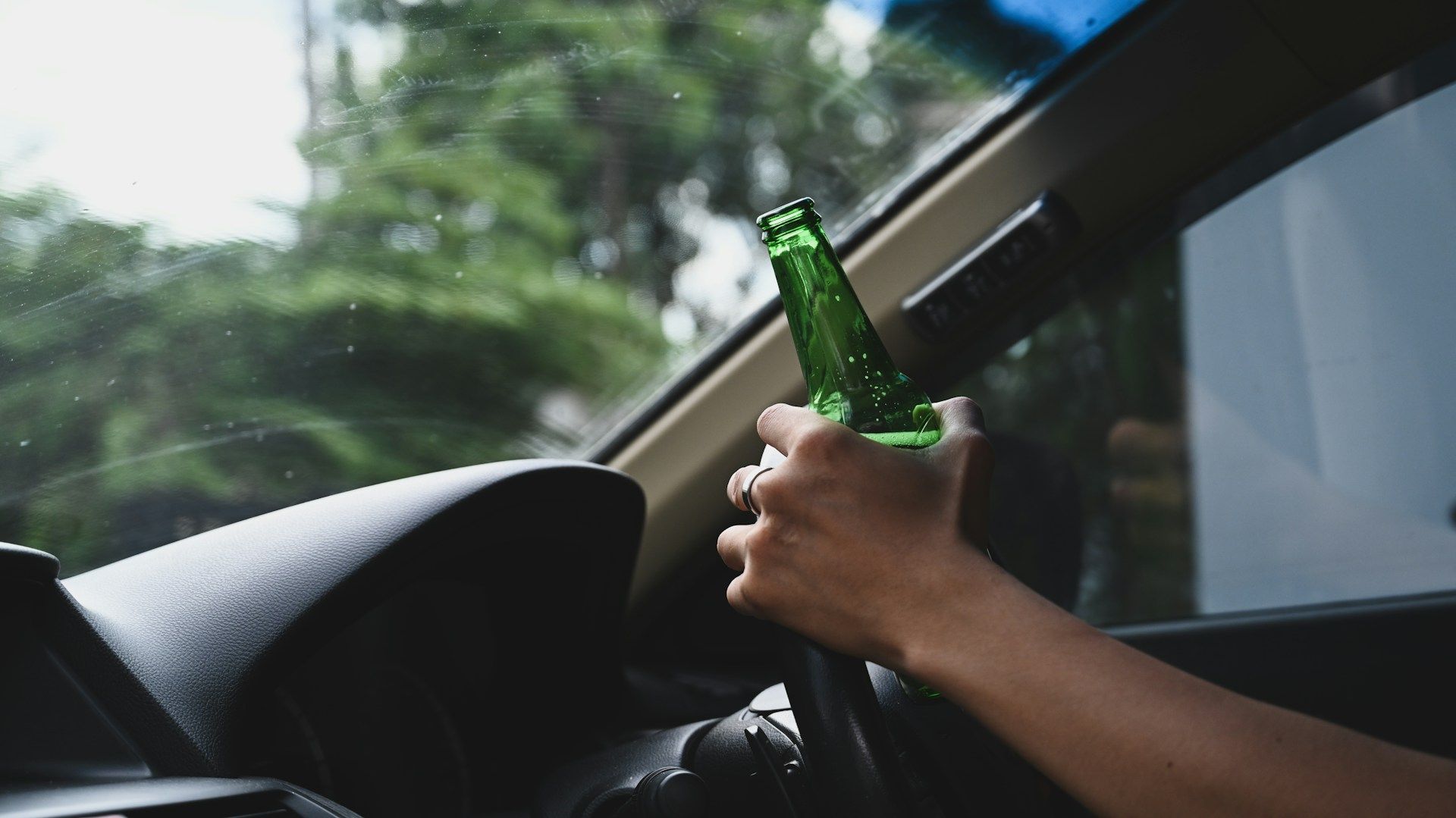 A person holding a green glass bottle while driving a vehicle.