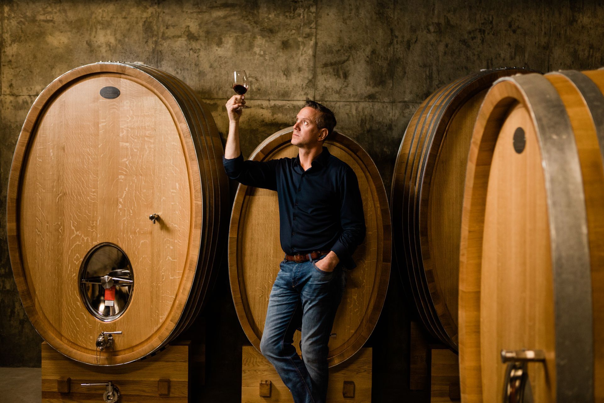 Man standing in a wine cellar beside large wooden barrels, holding a wine glass and looking upward