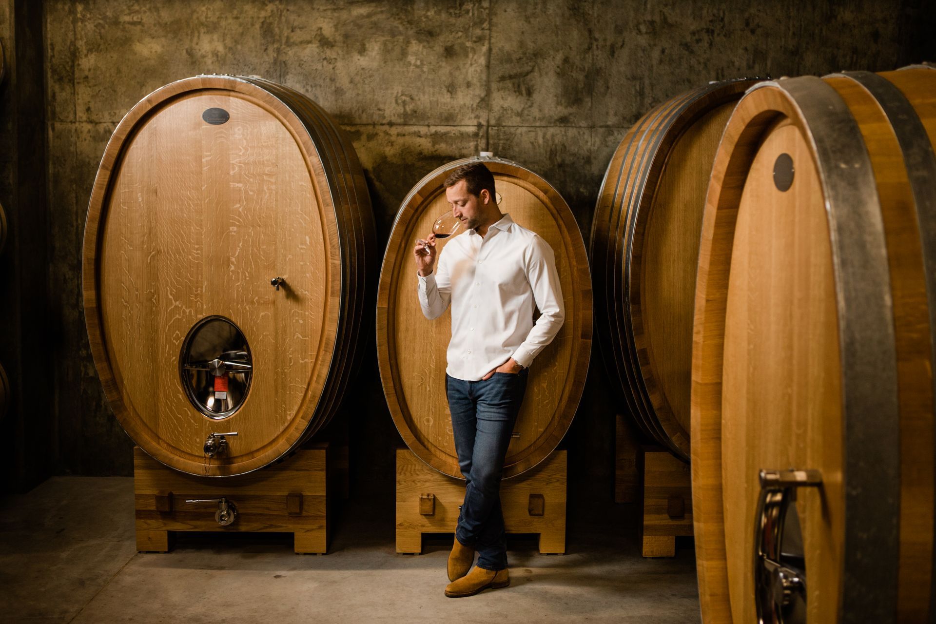 Person leaning against large wooden wine barrels in a dim wine cellar