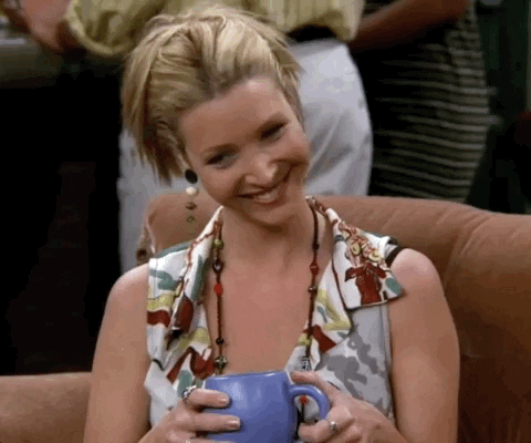 Phoebe from Friends holding a blue mug of coffee, wearing a patterned top in a crowded indoor setting