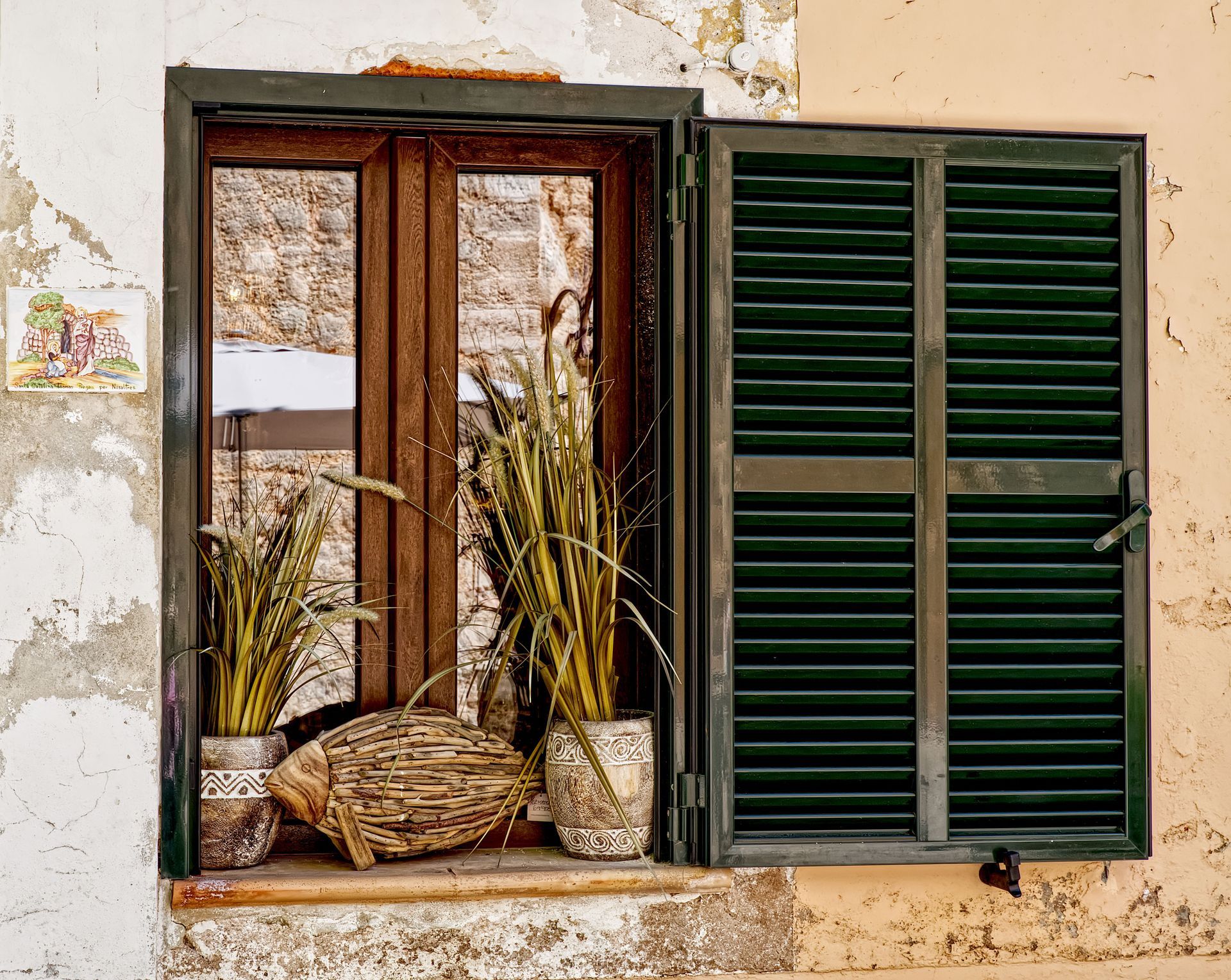Old stucco wall with an open wooden window, green shutters, and potted plants on the sill