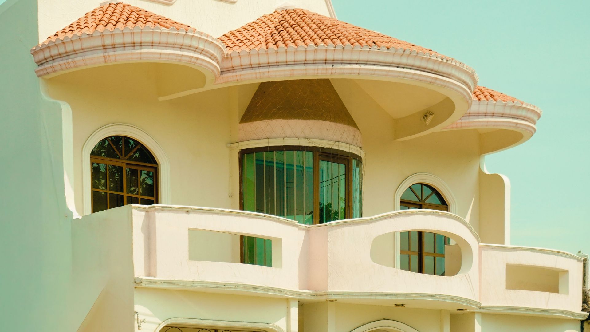 Cream-colored house with curved balconies, green windows, and a terracotta-tiled roof under bright sunlight