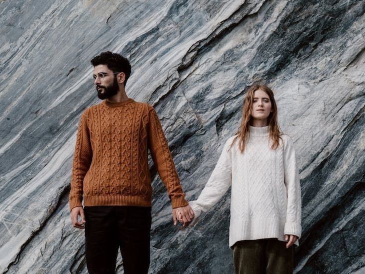 a man and a woman holding hands in front of a rock wall wearing chunky knitted jumpers