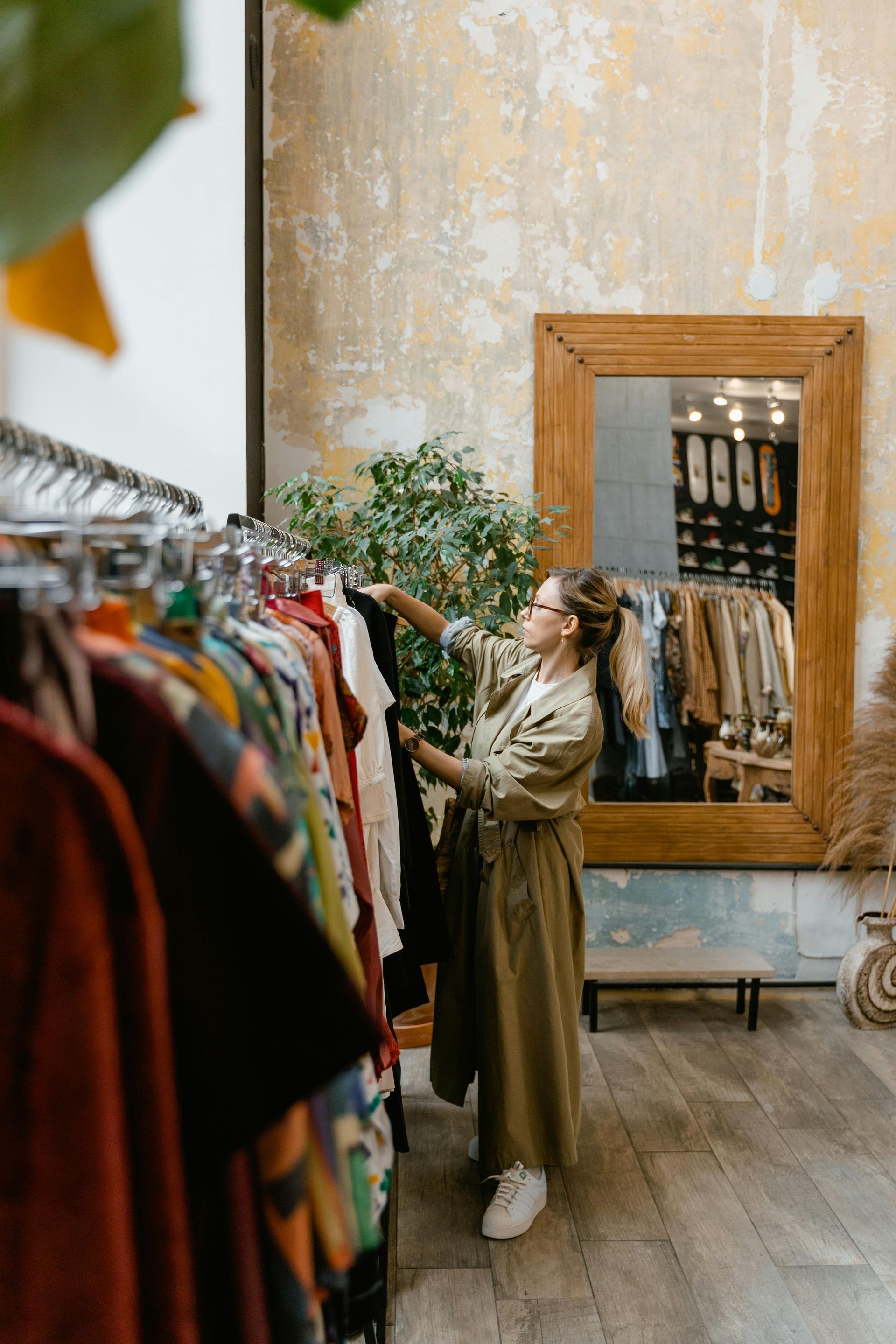 Woman browsing clothes on racks in a boutique with a large mirror and worn beige walls