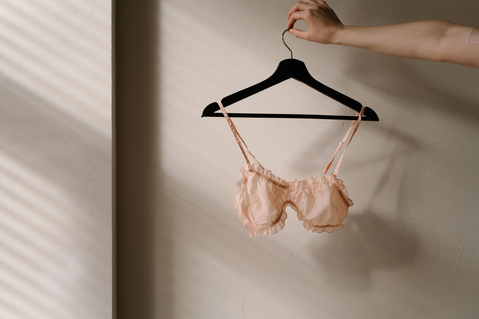 A hand holding a black hanger with a peach lace bra against a beige wall.