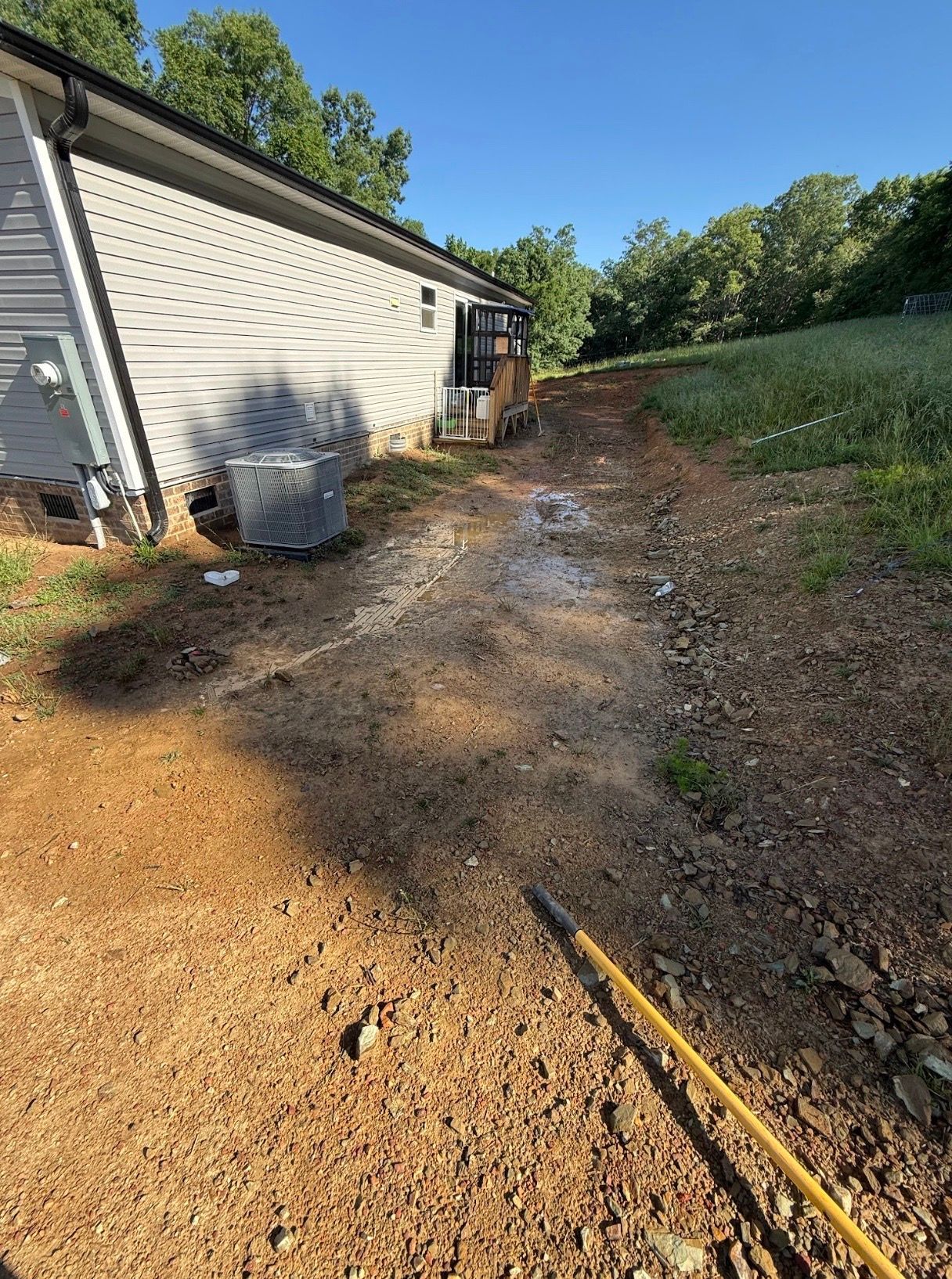 Gravel driveway beside a house, with a dirt path, trash bin, and hedge in bright sunlight.