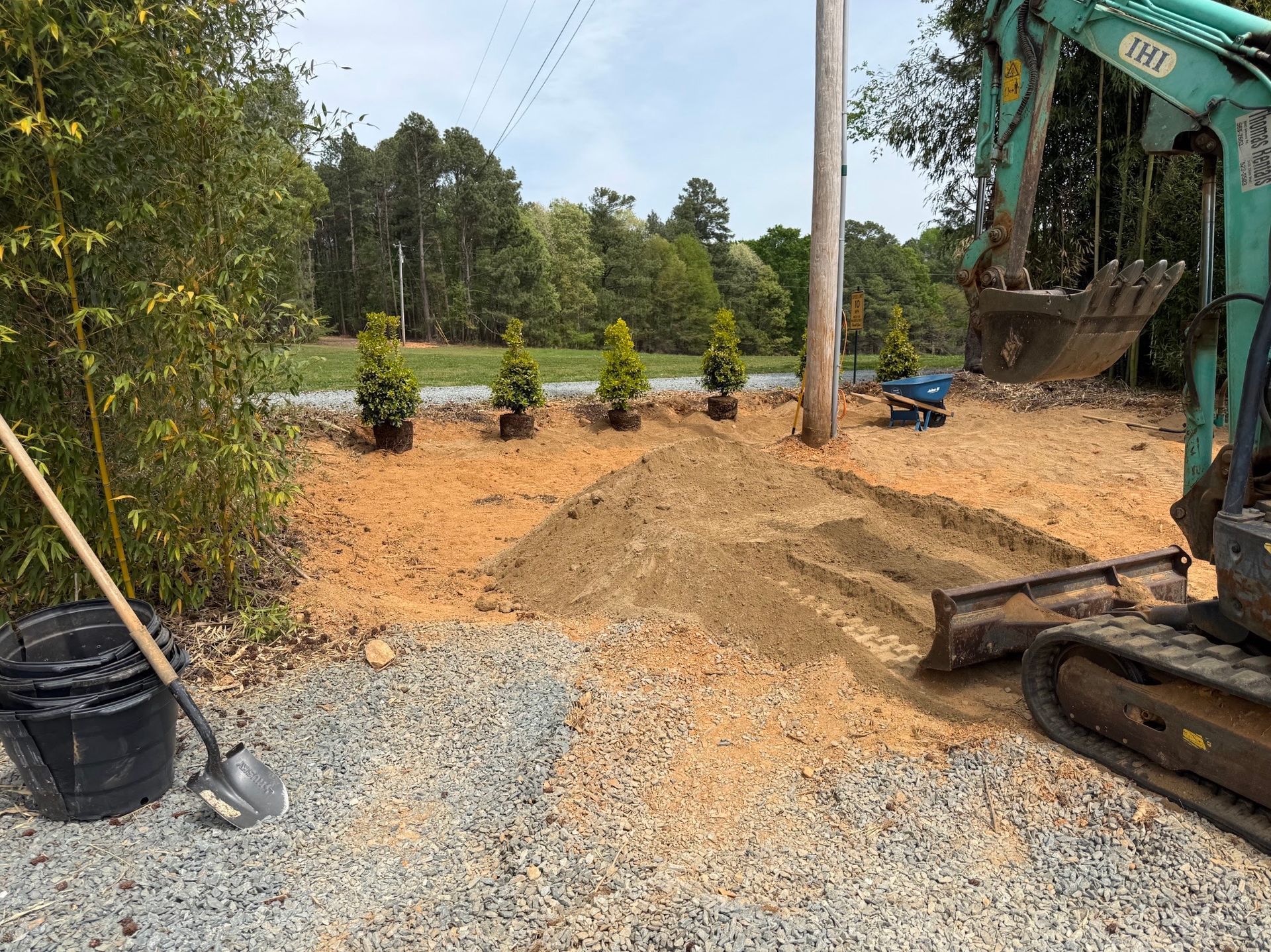 Construction site with excavator, gravel driveway, dirt piles, and roadside greenery