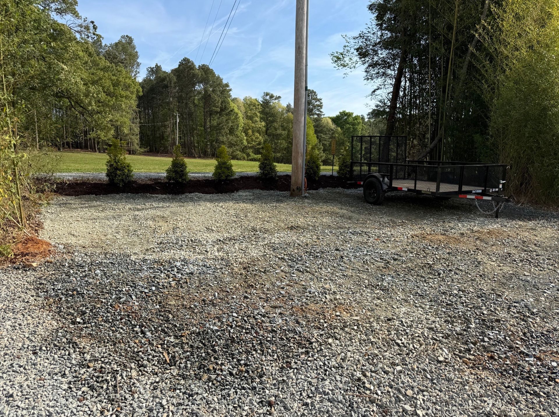 Gravel driveway beside trees and a utility pole, with a flatbed trailer in the background.