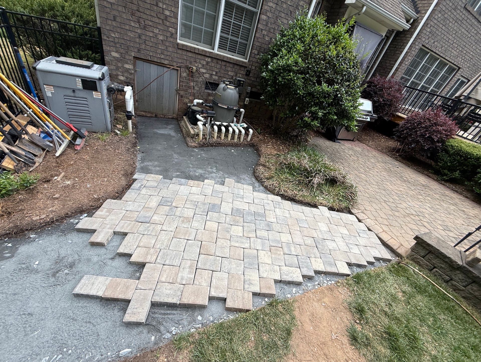 Partially paved backyard patio with paver stones around a small brick house and garden beds